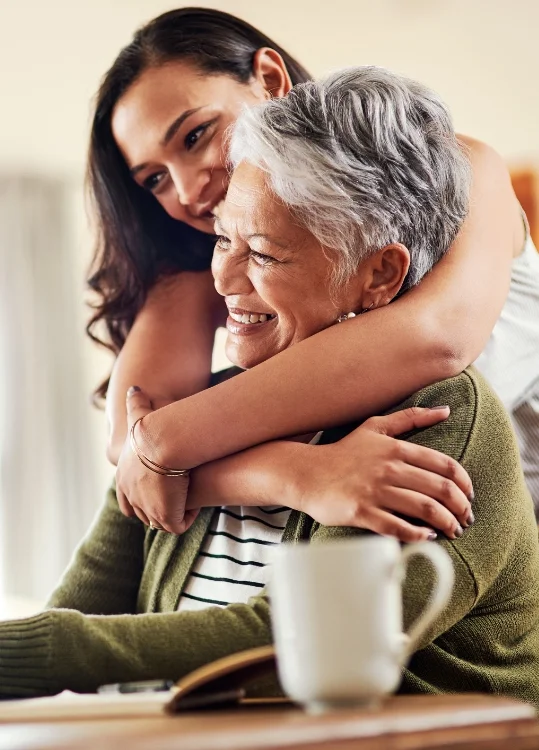adult daughter hugs senior mother from behind while she's seated at a table
