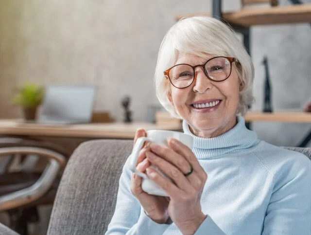 senior woman seated on cozy chair smiles and holds coffee mug