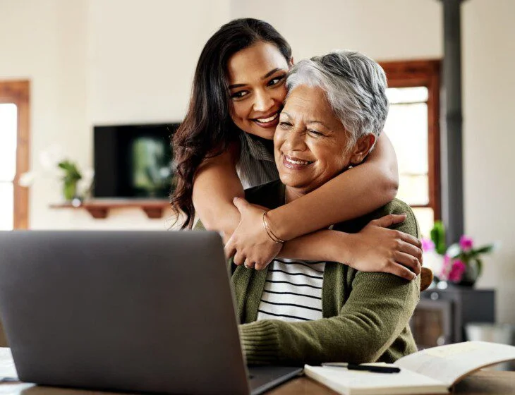 senior woman sits at laptop smiling while hugged from behind by her adult daughter