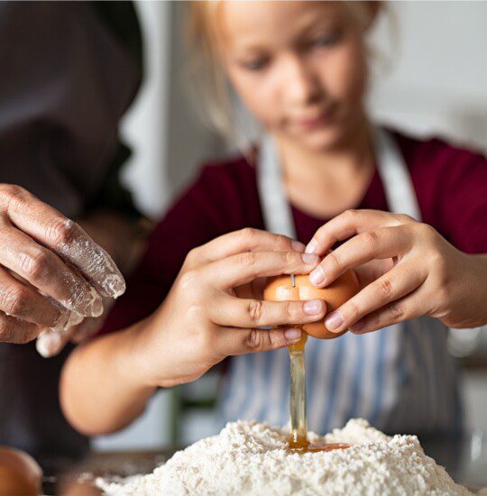 grandparent shows granddaughter how to crack egg while making bread