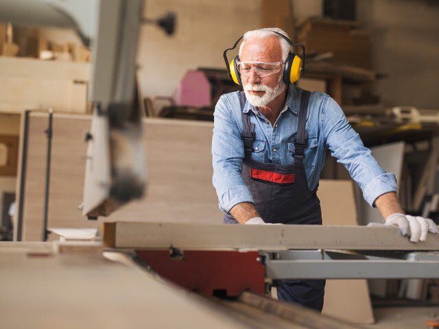 senior man wearing protective glasses and earmuffs while sawing piece of wood in his workshop