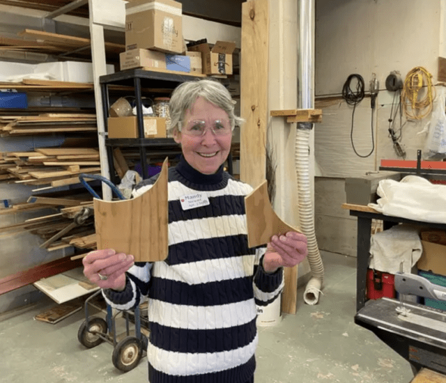 Oak Trace Senior Living resident Mandy Stewart smiles inside a workshop, wearing protective goggles and holding up pieces of wood