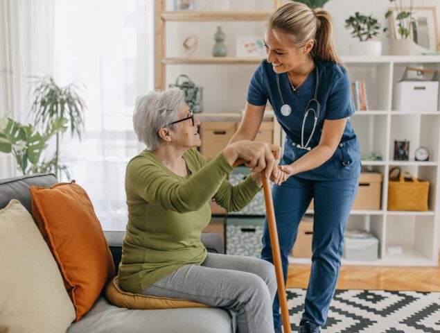 caregiver smiles and assists senior woman seated on a couch in her apartment