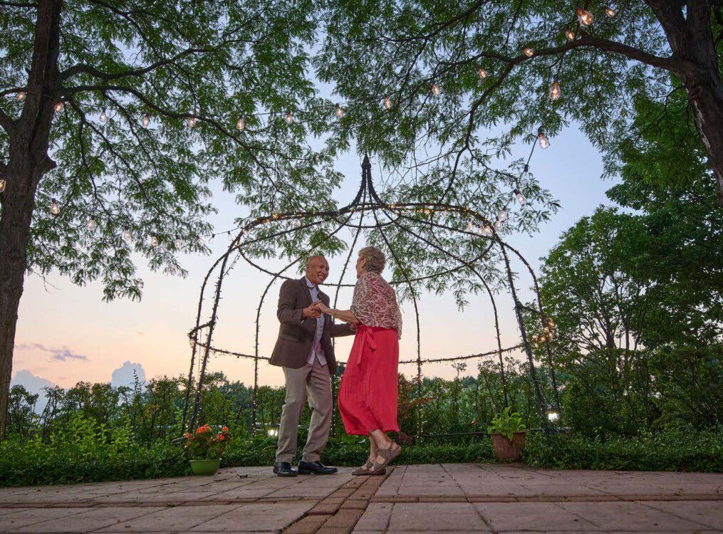 senior couple in elegant clothing dances together outside at sunset under fairy lights at Oak Trace Senior Living Community