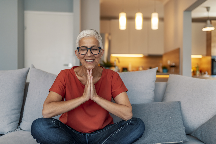 senior woman sits with her legs up, practicing meditation in her apartment at Oak Trace Senior Living Community