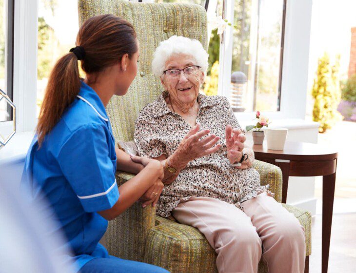 Senior woman seated in chair smiles and talks to her caretaker, who kneels beside her