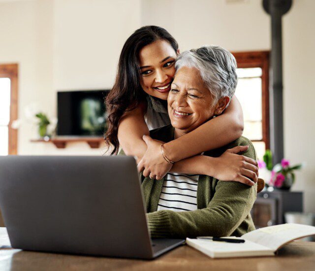 senior woman sits at laptop smiling while hugged from behind by her adult daughter