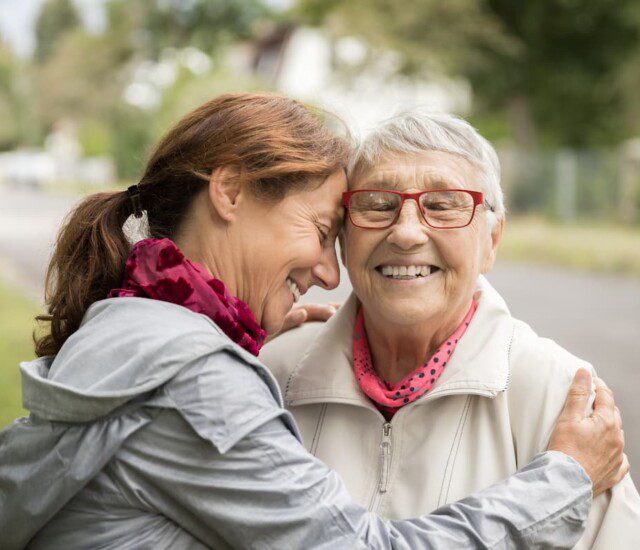 Senior woman and her adult daughter embrace for a hug on a walking path outside of Oak Trace Senior Living Community