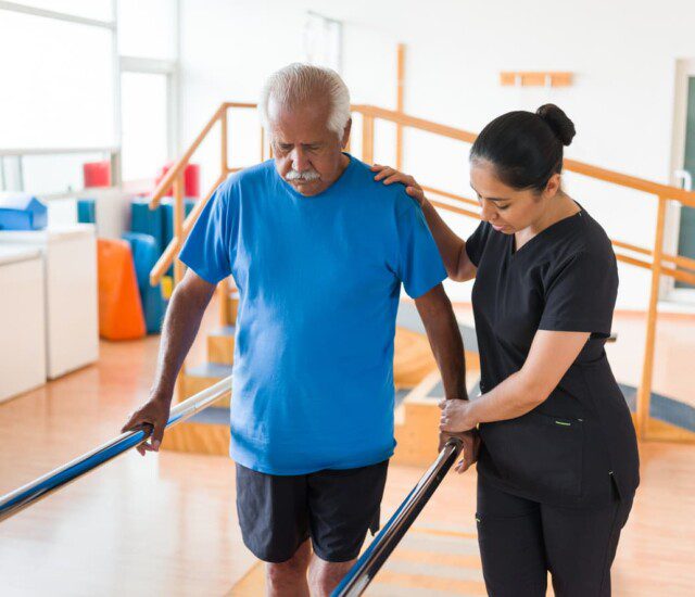 senior man uses bars to hold himself up with the aid of a physical therapist during a rehabilitation session