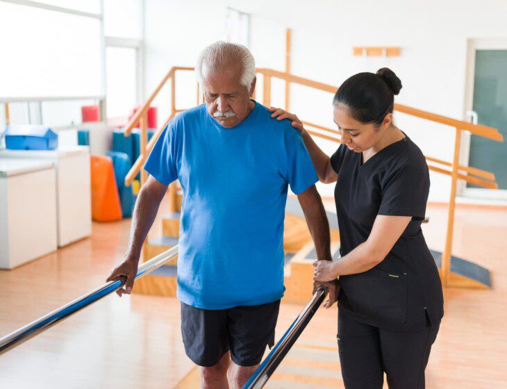 senior man uses bars to hold himself up with the aid of a physical therapist during a rehabilitation session